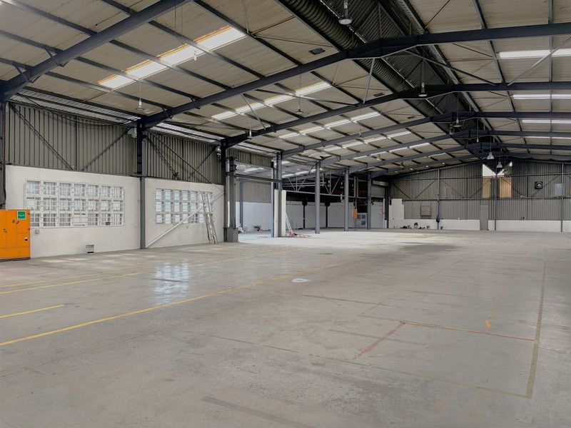 A spacious industrial warehouse featuring high ceilings with exposed steel trusses, concrete flooring, and natural light provided by integrated skylights.