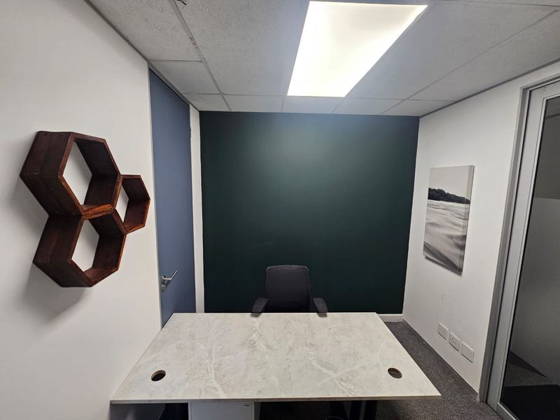 A compact, modern office space featuring a built-in desk, dark accent wall, carpeted flooring, and a glass partition door.