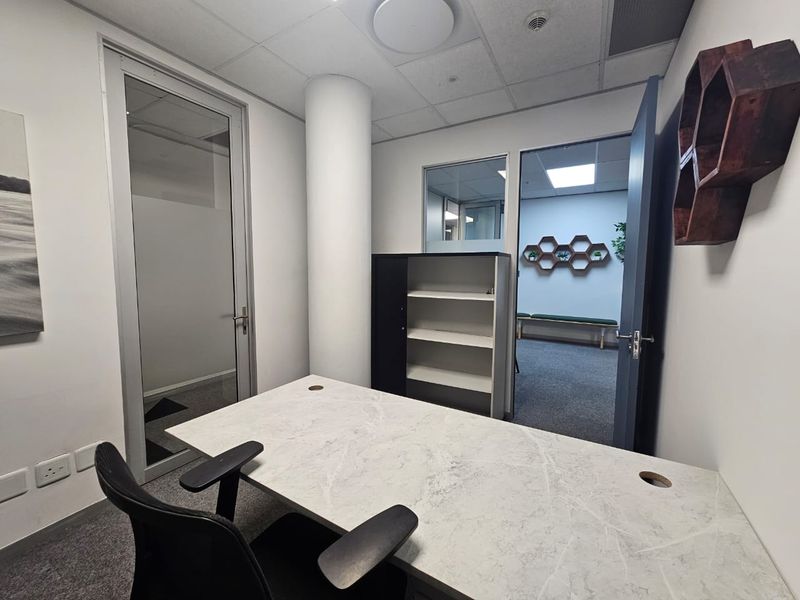 A professional office space featuring a modern desk, carpeted flooring, a structural column, and glass partition doors providing a view into an adjacent room.