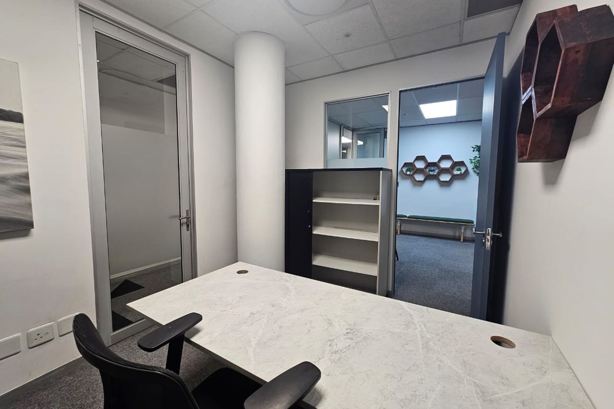 A professional office space featuring a modern desk, carpeted flooring, a structural column, and glass partition doors providing a view into an adjacent room.