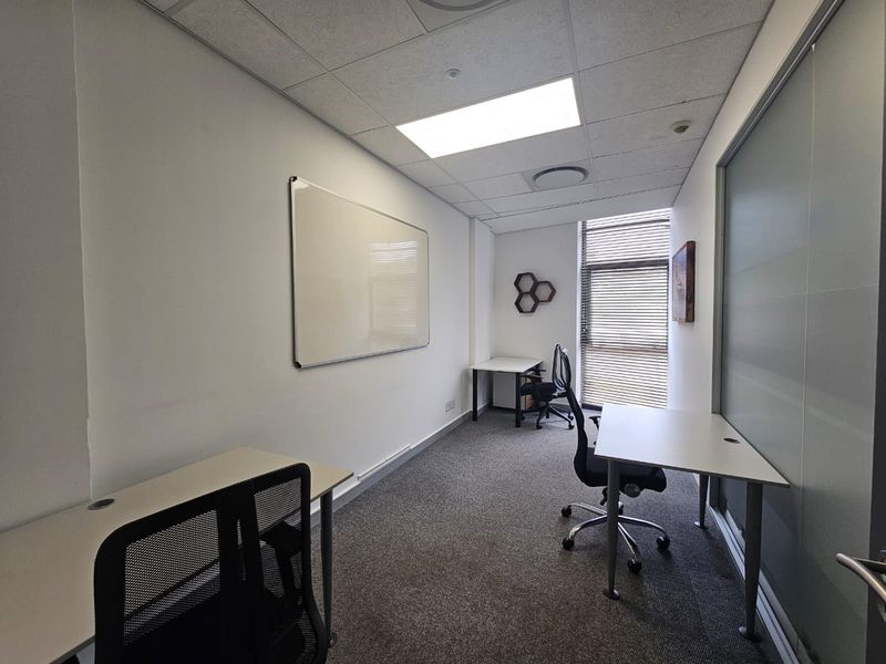 A functional office space featuring carpeted flooring, a suspended ceiling with recessed lighting, a frosted glass partition, and a wall-mounted whiteboard.