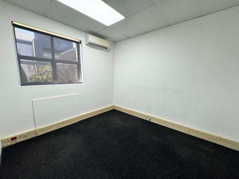 A vacant office space featuring dark carpeting, a suspended ceiling with recessed lighting, and a wall-mounted split-system air conditioning unit.