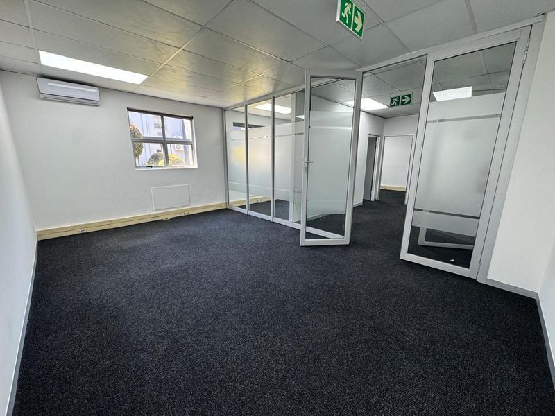 A clean, vacant office space featuring modern glass partitions, dark carpeting, recessed lighting, and a wall-mounted air conditioning unit.