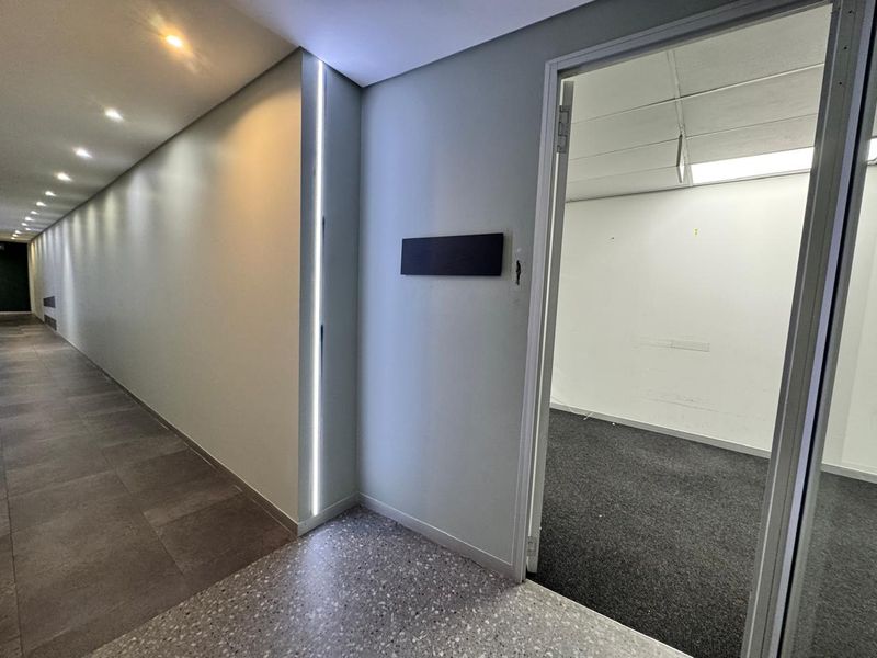 A modern office hallway featuring recessed LED strip lighting, tiled flooring, and an open doorway leading into an empty carpeted office space.