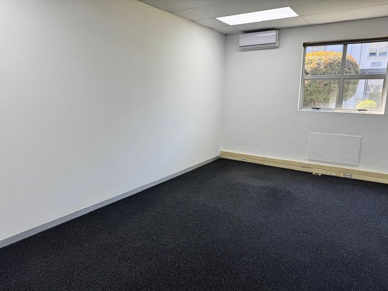 A vacant office space featuring dark carpeting, a suspended ceiling with recessed lighting, and a wall-mounted air conditioning unit.