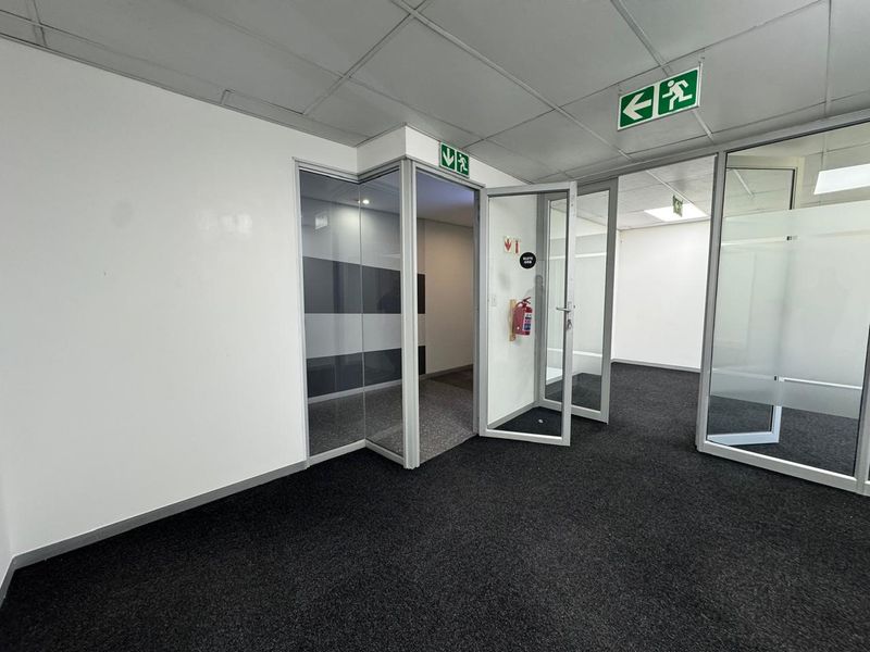 A vacant office space featuring glass partitions, suspended ceilings, and carpeted flooring, equipped with emergency exit signage and fire safety equipment.