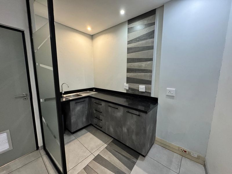 A modern, compact office kitchenette featuring dark grey cabinetry, granite countertops, a stainless steel sink, and decorative tiled backsplash.