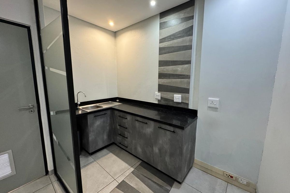 A modern, compact office kitchenette featuring dark grey cabinetry, granite countertops, a stainless steel sink, and decorative tiled backsplash.