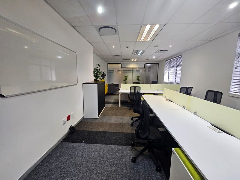A professional open-plan office space featuring modular desk workstations, carpeted flooring, suspended ceilings with recessed lighting, and large windows with fitted blinds.