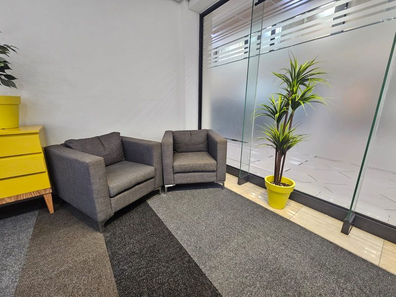 A modern office waiting area featuring a glass partition wall, neutral carpet flooring, and clean white walls.