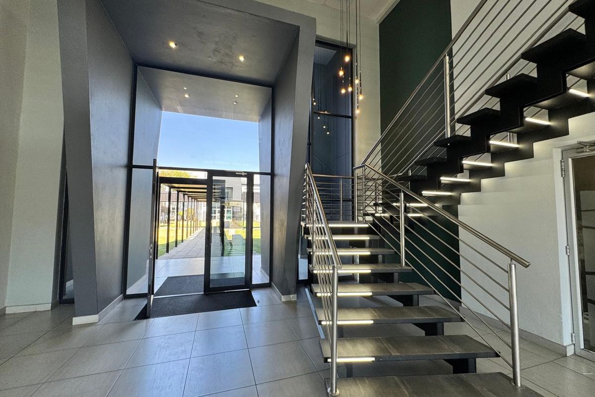 Modern office entrance foyer featuring a striking staircase with integrated lighting, stainless steel balustrades, and large glass doors leading to an outdoor walkway.