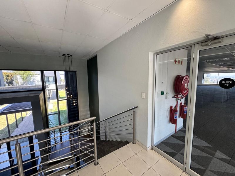 A bright office hallway featuring a stainless steel staircase balustrade, tiled flooring, and integrated fire safety equipment leading into Suite F6.