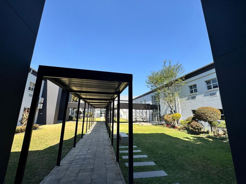 A well-maintained office courtyard featuring a covered walkway, paved pathways, and landscaped garden areas between the building wings.
