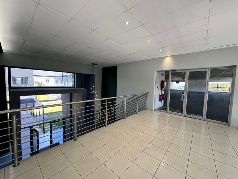A spacious, tiled office hallway featuring a stainless steel balustrade, suspended ceiling with recessed lighting, and glass partition doors leading to adjacent office areas.