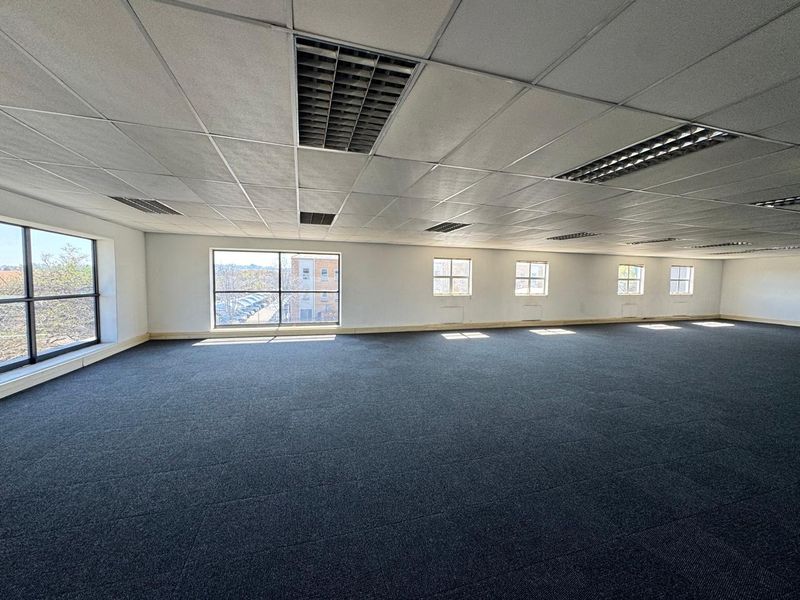 A spacious, open-plan office area featuring carpet tiles, a suspended ceiling with recessed lighting, and large windows providing ample natural light.