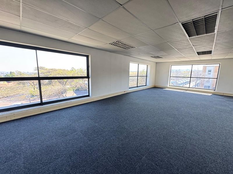 A bright, open-plan office space featuring carpet tiles, a suspended ceiling with recessed lighting, and large windows providing ample natural light.