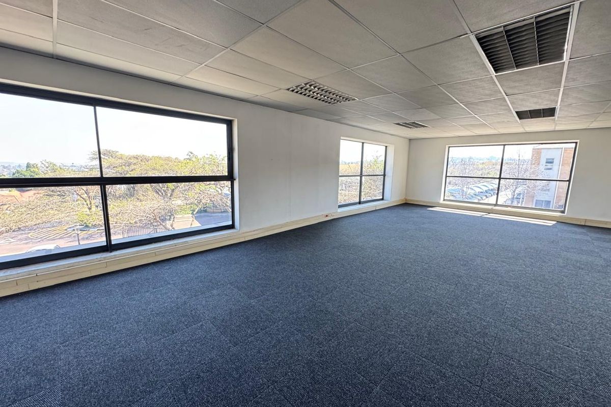A bright, open-plan office space featuring carpet tiles, a suspended ceiling with recessed lighting, and large windows providing ample natural light.