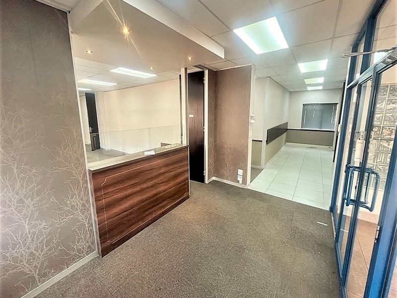A vacant office reception area featuring a built-in wooden desk, suspended ceiling with recessed lighting, and a mix of carpet and tiled flooring.