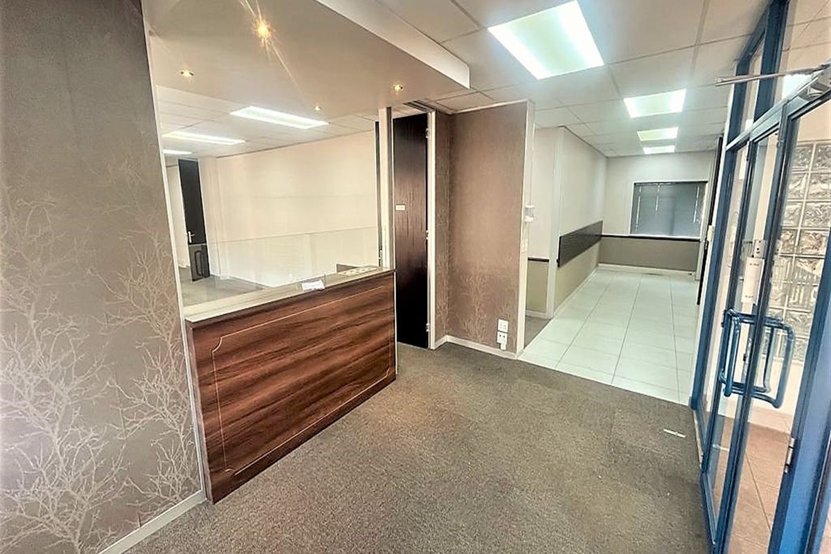 A vacant office reception area featuring a built-in wooden desk, suspended ceiling with recessed lighting, and a mix of carpet and tiled flooring.