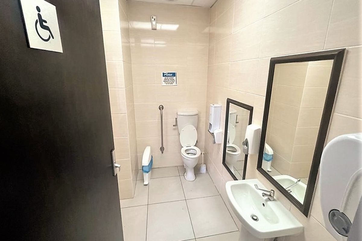 A clean, functional accessible office bathroom featuring a pedestal sink, toilet with grab rail, and neutral wall tiling.