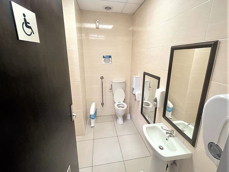 A clean, functional accessible office bathroom featuring a pedestal sink, toilet with grab rail, and neutral wall tiling.