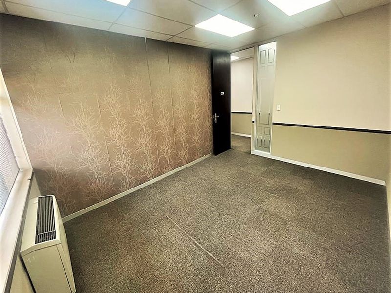 A vacant office space featuring carpeted flooring, a suspended ceiling with recessed lighting, and a wall-mounted HVAC unit.