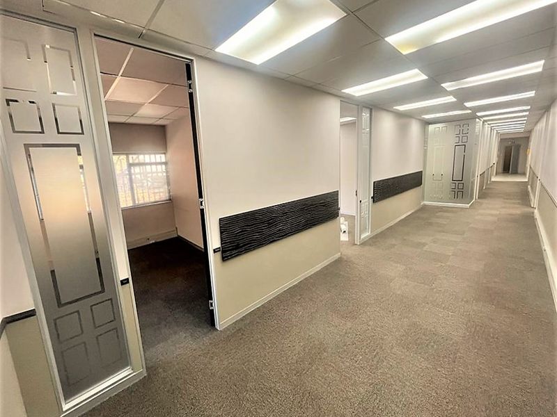 A clean, well-lit office hallway featuring carpeted floors, suspended ceilings, and frosted glass doors leading into individual office spaces.