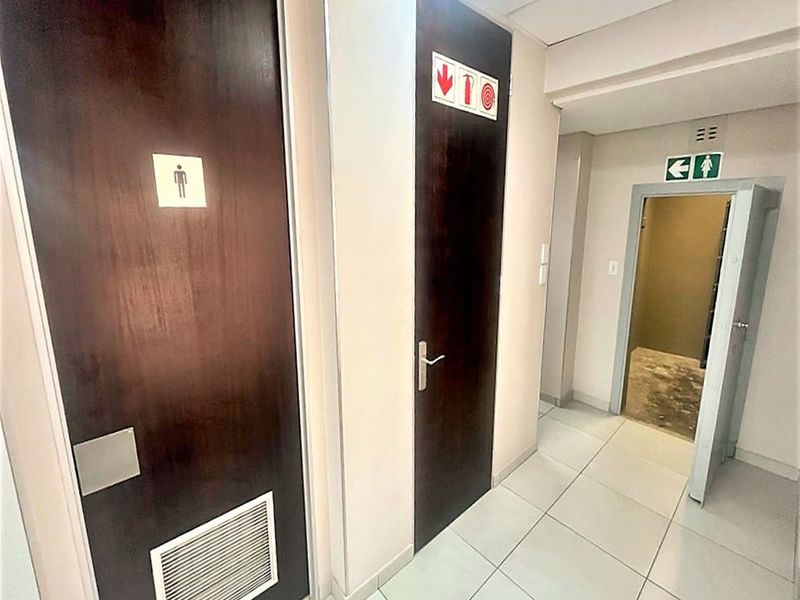 A functional office hallway featuring tiled flooring, multiple access doors with signage, and a drop ceiling, providing access to restroom facilities.