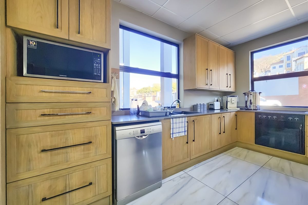 A well-maintained office kitchen featuring light wood cabinetry, integrated appliances, modern tiled flooring, and ample natural light from large windows.