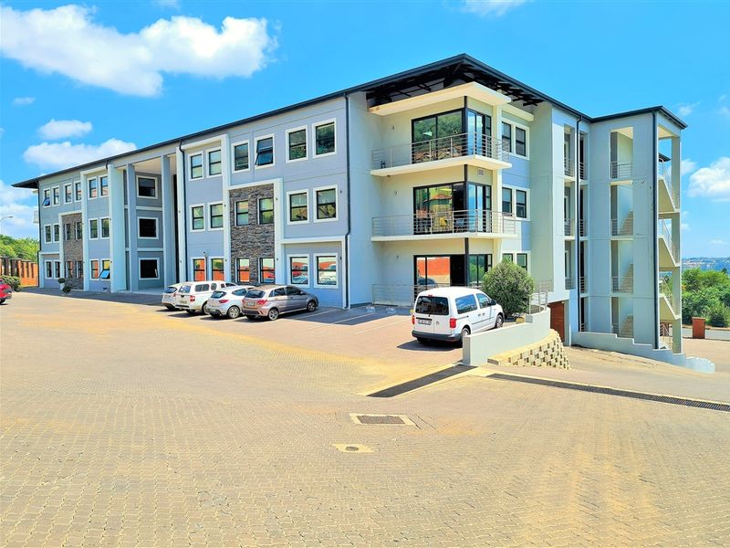 Modern multi-story office building featuring stone cladding accents, private balconies with metal railings, and an expansive paved parking area.
