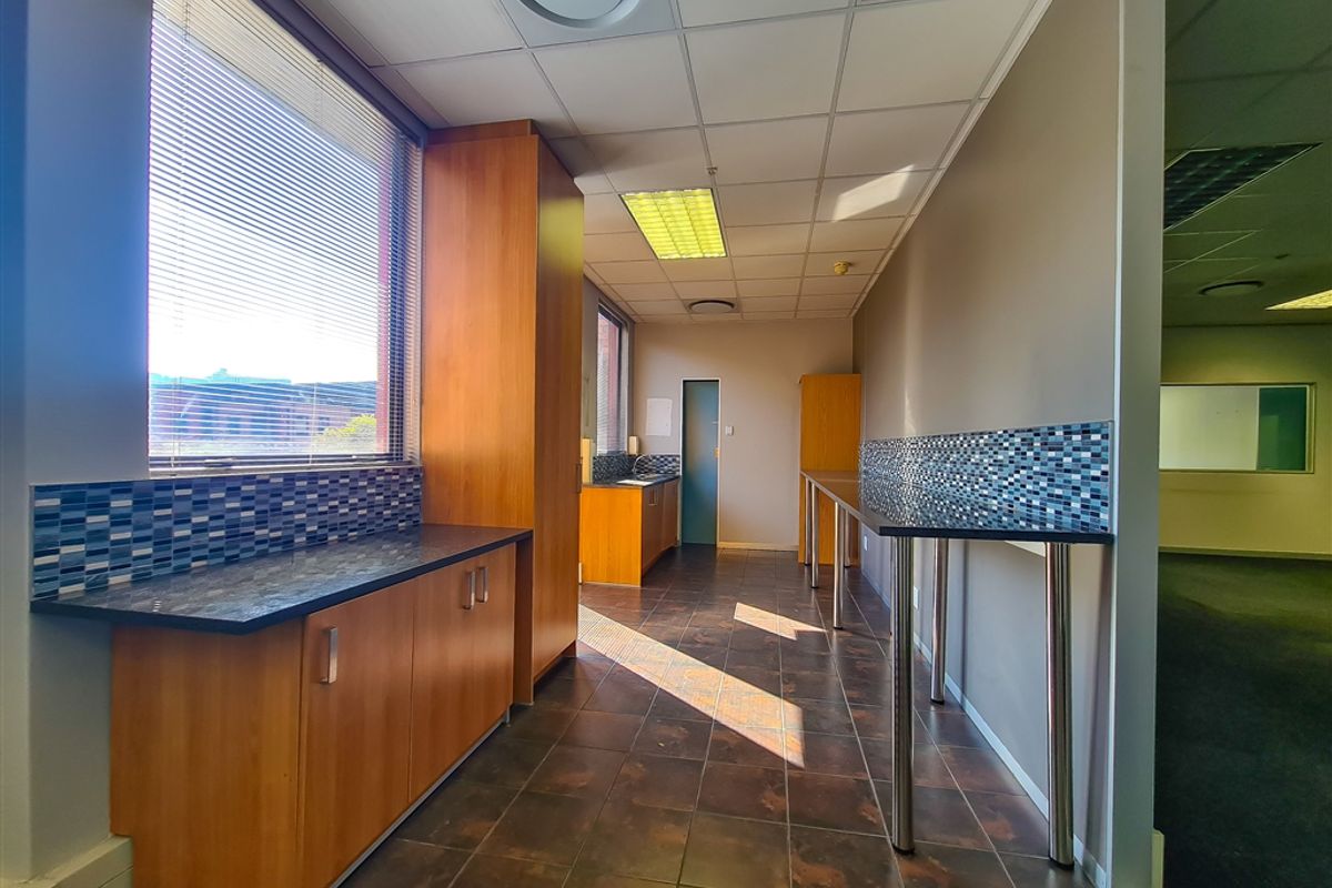 A clean, functional office kitchenette featuring wood-finish cabinetry, dark countertops, mosaic tile backsplashes, and durable ceramic floor tiles.