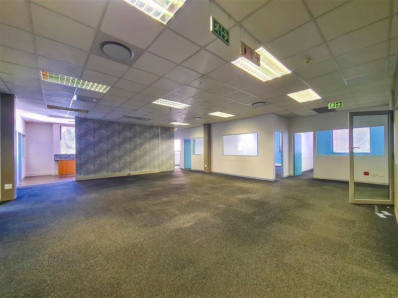 Spacious open-plan office area featuring suspended ceilings, recessed lighting, carpeted flooring, and integrated glass partitions with a small kitchenette corner.