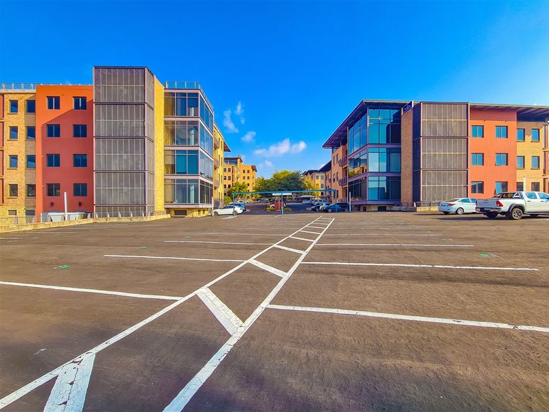 A wide-angle view of a modern office complex featuring a large asphalt parking area between two multi-story buildings with glass facades.