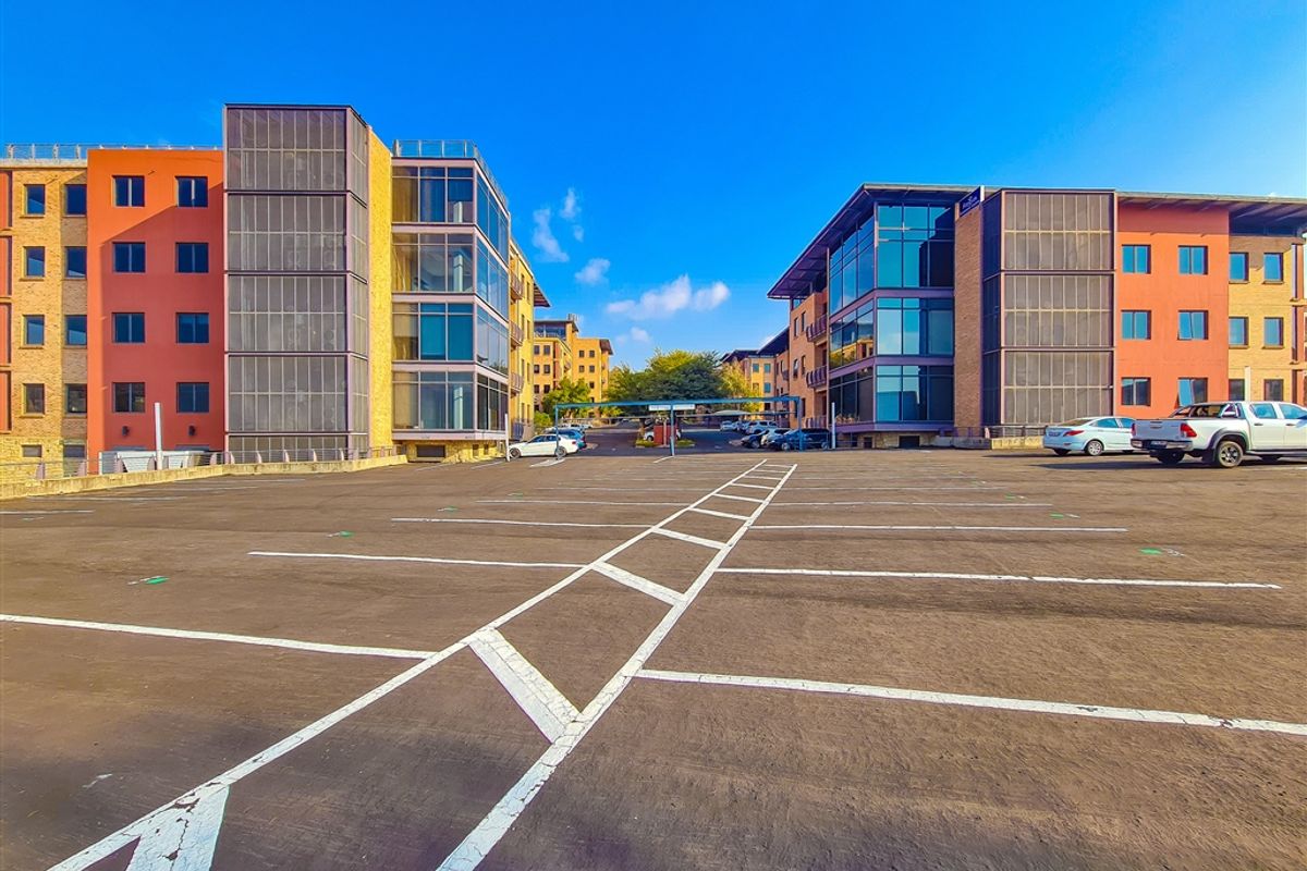 A wide-angle view of a modern office complex featuring a large asphalt parking area between two multi-story buildings with glass facades.