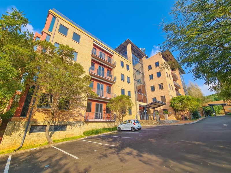 A multi-story office building featuring a brick facade, glass curtain walling, and private balconies, set within a landscaped office park with dedicated parking.