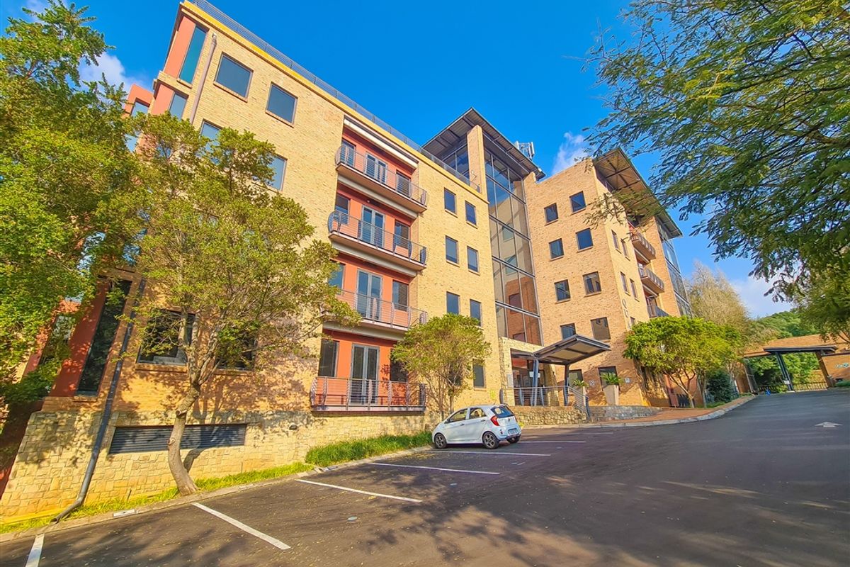 A multi-story office building featuring a brick facade, glass curtain walling, and private balconies, set within a landscaped office park with dedicated parking.