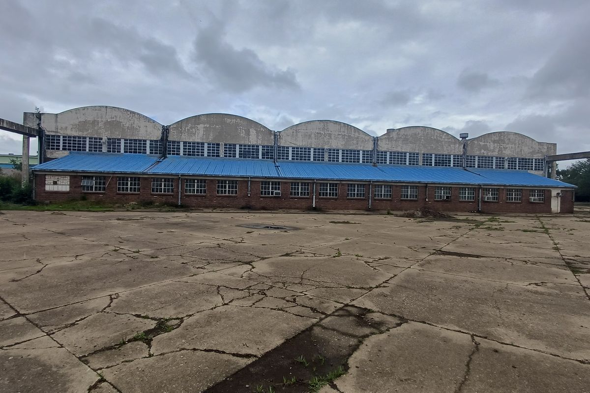 Large industrial warehouse facility featuring a distinctive arched roof design, brick facade, and extensive concrete yard area requiring maintenance.