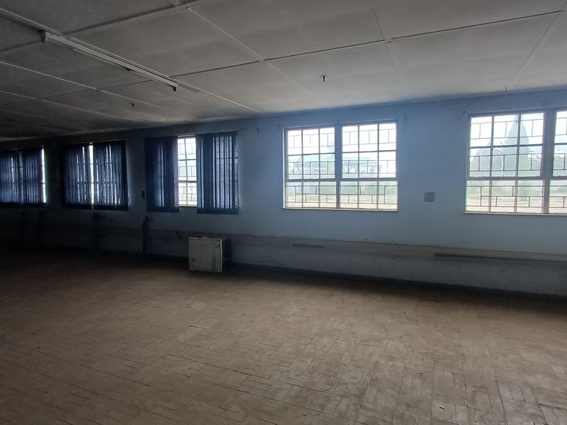 A large, vacant industrial office or utility space featuring multiple windows, tiled flooring, and a suspended ceiling, requiring significant refurbishment.