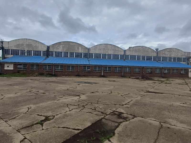 Large industrial warehouse facility featuring a distinctive arched roof design, brick facade, and extensive concrete yard area requiring maintenance.