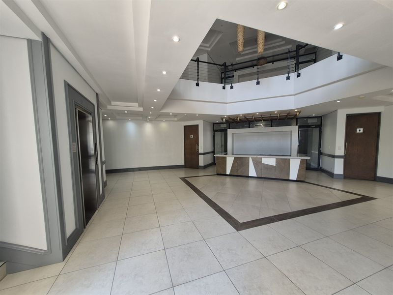 Spacious office building entrance lobby featuring a reception desk, elevator access, tiled flooring, and a double-volume ceiling with mezzanine level.