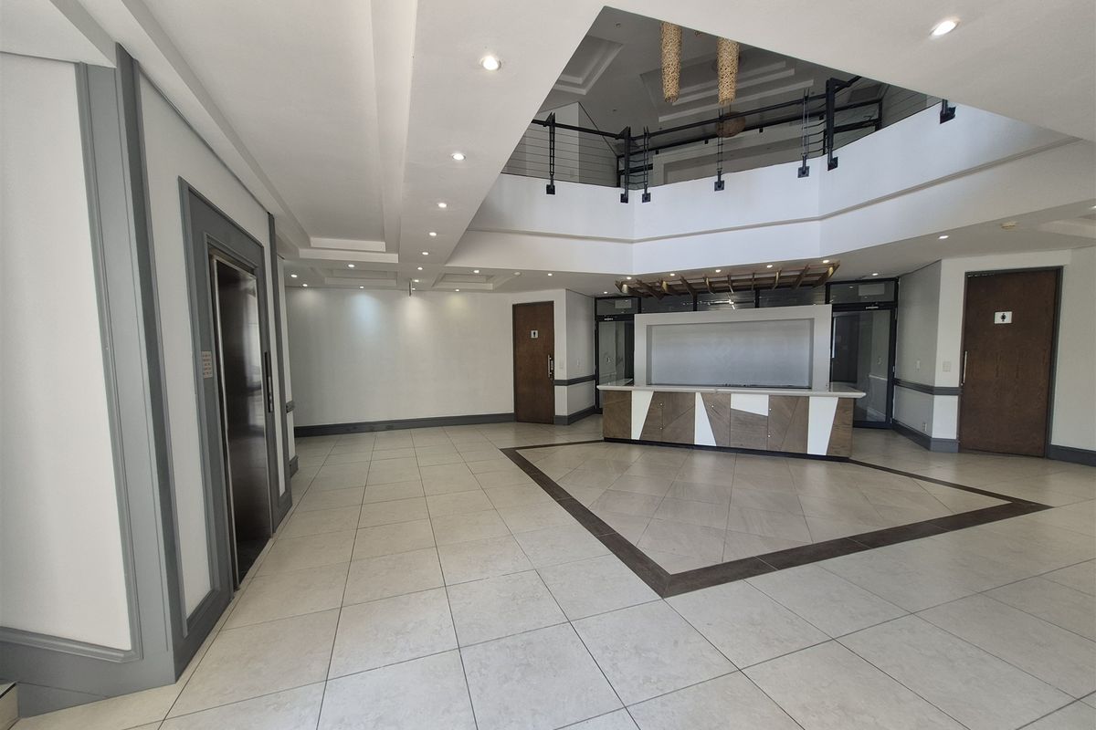 Spacious office building entrance lobby featuring a reception desk, elevator access, tiled flooring, and a double-volume ceiling with mezzanine level.
