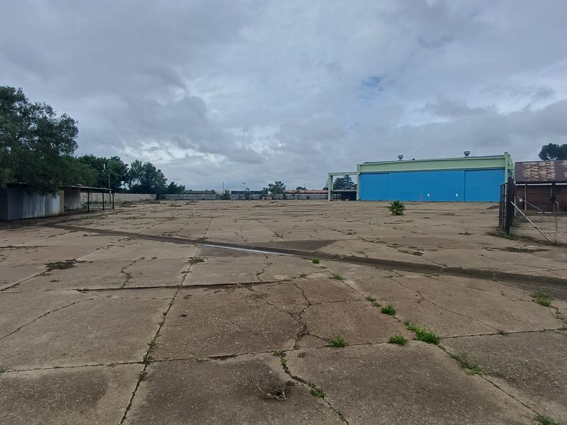 Expansive industrial yard featuring a large concrete paved area, surrounding outbuildings, and a prominent blue industrial hangar structure.