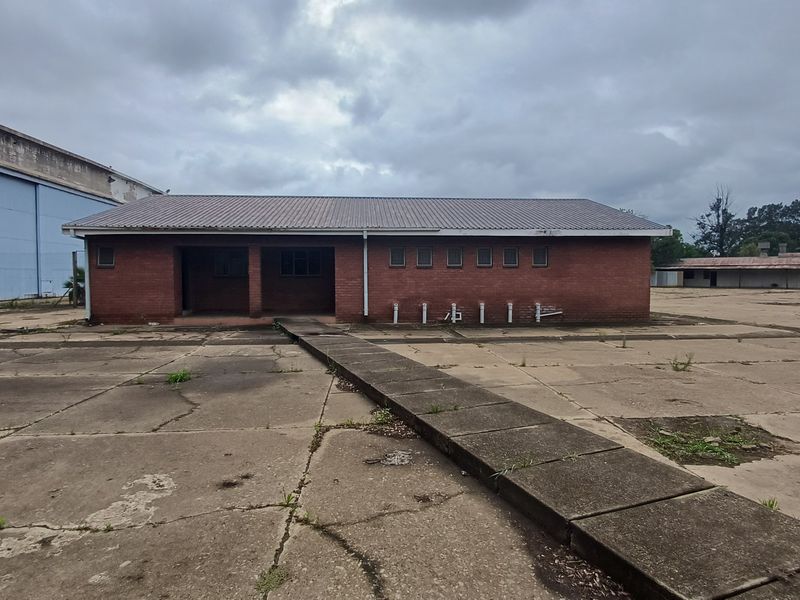 Exterior view of a single-story industrial brick building featuring a concrete apron and raised walkway, set within a large paved industrial yard.