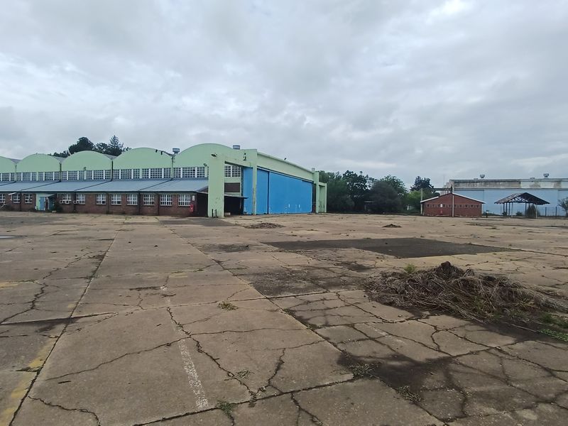 Large industrial hangar facility with extensive concrete yard space, featuring multiple arched roof sections and auxiliary outbuildings on a spacious site.