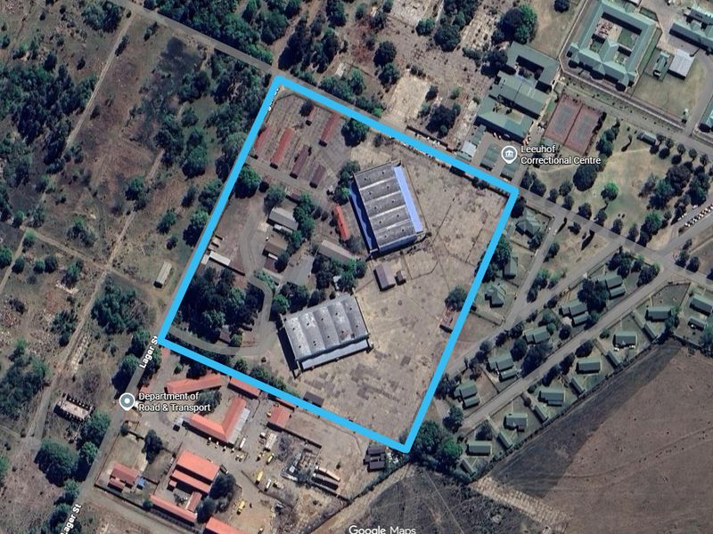 Aerial view of an industrial property featuring two large warehouse structures, paved access areas, and surrounding open land within a fenced perimeter.