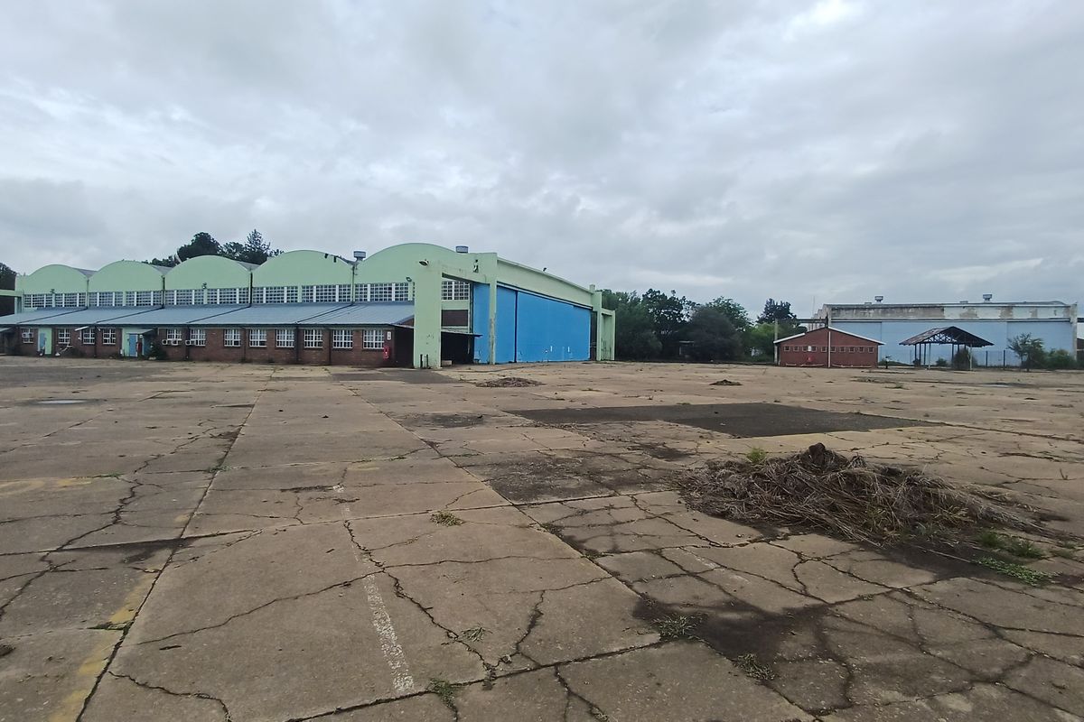 Large industrial hangar facility with extensive concrete yard space, featuring multiple arched roof sections and auxiliary outbuildings on a spacious site.