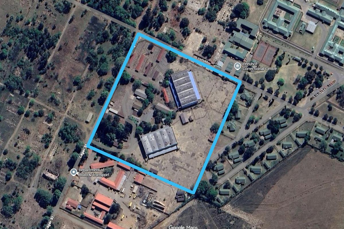 Aerial view of an industrial property featuring two large warehouse structures, paved access areas, and surrounding open land within a fenced perimeter.