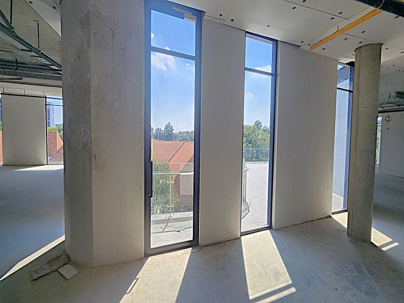 An unfinished open-plan office space featuring exposed concrete columns, high ceilings, and large windows providing access to a balcony with glass balustrades.