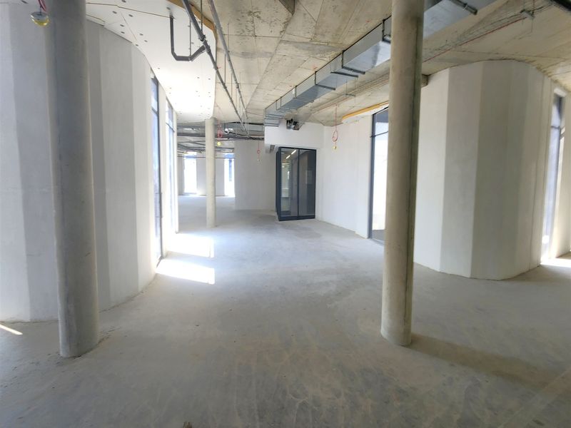 An open-plan office space in shell condition, featuring exposed concrete ceilings, structural columns, and large windows awaiting final fit-out and finishes.