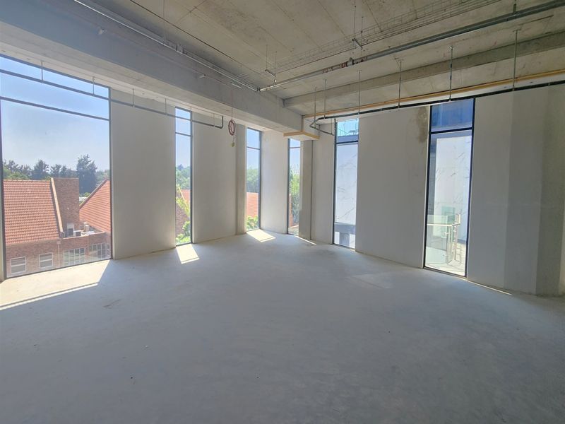 An open-plan office space in shell condition featuring high exposed concrete ceilings, large windows providing natural light, and access to a balcony.
